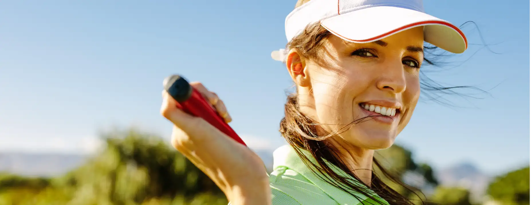 A woman wearing a white visor with red trim and holding a red golf club.